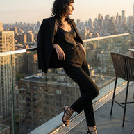 Woman in black suit and heels poses on rooftop balcony with city skyline at sunset.