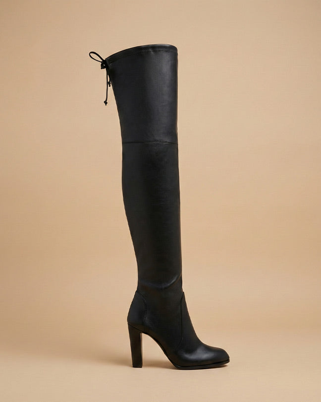 Black over-the-knee high heel boot with tie at back, displayed on beige background