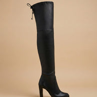 Black over-the-knee high heel boot with tie at back, displayed on beige background
