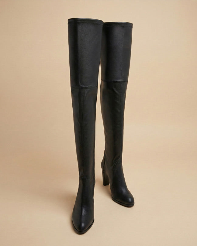 Black over-the-knee heeled boots with pointed toes on beige background