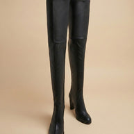 Black over-the-knee heeled boots with pointed toes on beige background