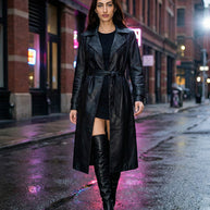 Woman wearing black leather trench coat and knee high boots walking on city street at night