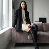 Woman in a black blazer, cream sweater dress, and over-the-knee boots sitting on a sofa