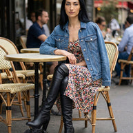 Woman in floral dress, denim jacket, and black knee-high boots sitting at outdoor café