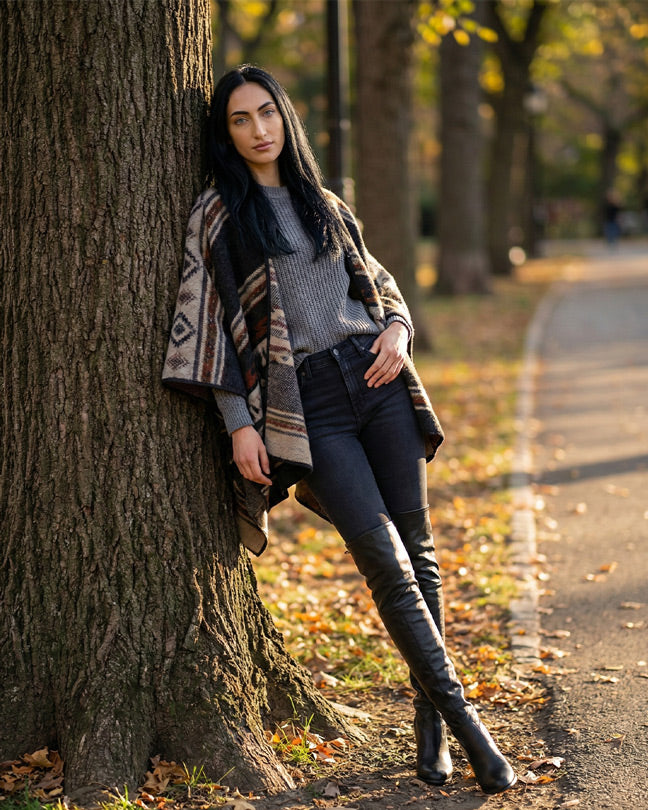 Woman in patterned poncho, gray sweater, black jeans, and knee-high boots leaning on tree in park