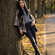Woman in patterned poncho, gray sweater, black jeans, and knee-high boots leaning on tree in park