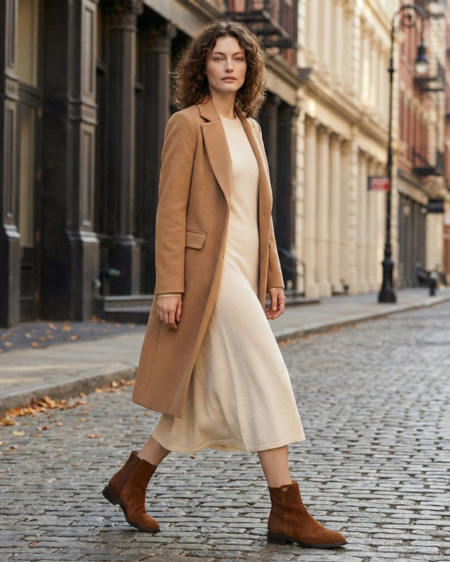 Woman in tan wool coat, cream midi dress, and brown suede boots walking on city street