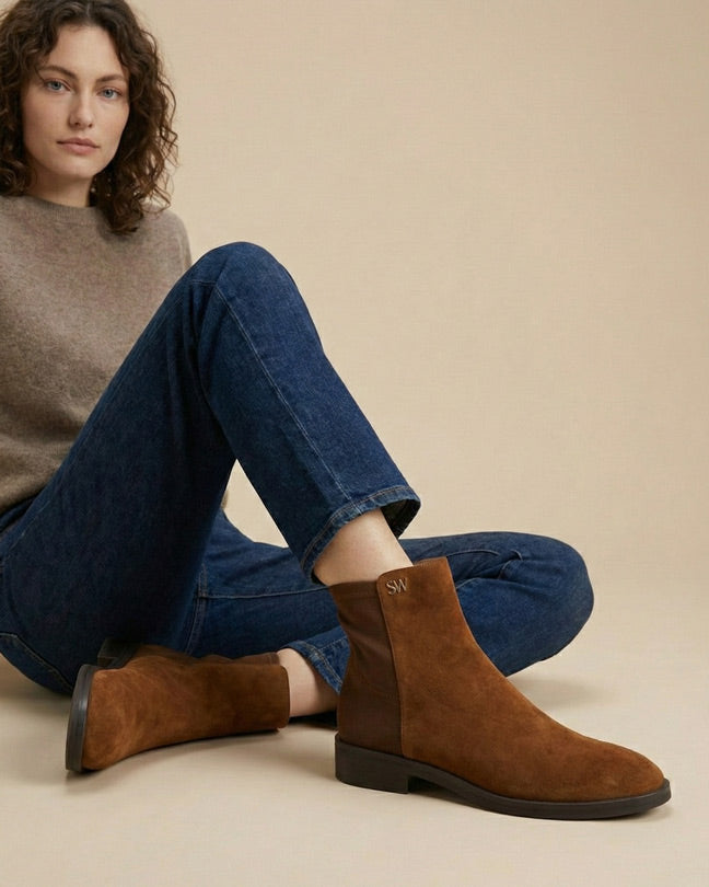 Woman wearing brown suede ankle boots, blue jeans, and a beige sweater, sitting casually