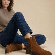Woman wearing brown suede ankle boots, blue jeans, and a beige sweater, sitting casually
