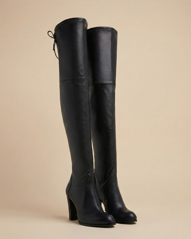 Black over-the-knee high heel boots with tie-back detail on neutral background