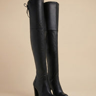 Black over-the-knee high heel boots with tie-back detail on neutral background