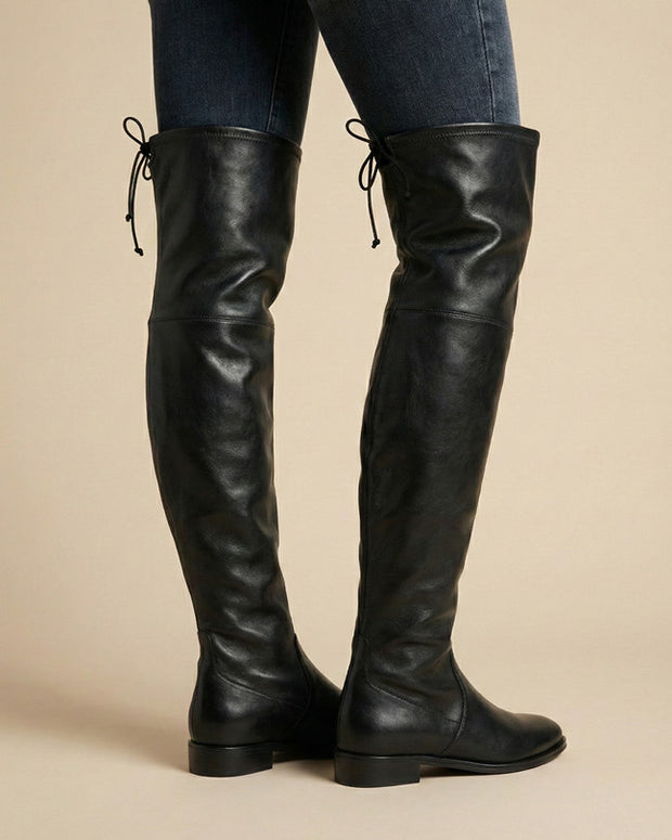 Black over-the-knee leather boots with low heels and lace-up back details, worn with jeans