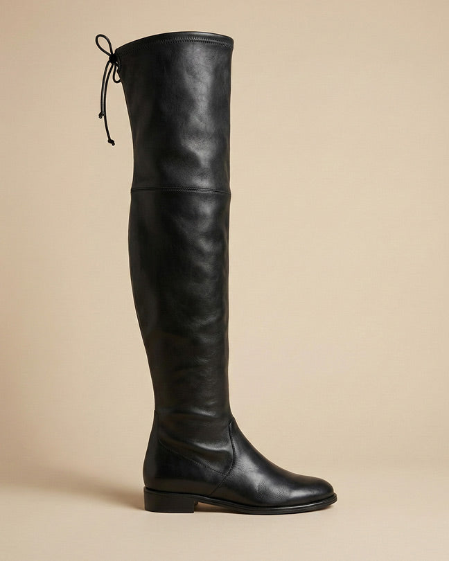 Black over-the-knee leather boot with flat heel and tie at top, side view on plain background