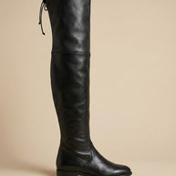Black over-the-knee leather boot with flat heel and tie at top, side view on plain background