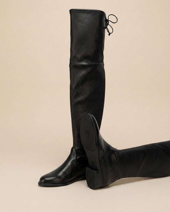 Black leather over-the-knee boots with low heel and tie detail, displayed on beige background
