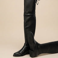 Black leather over-the-knee boots with low heel and tie detail, displayed on beige background