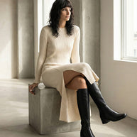 Woman in ribbed cream knit dress and black knee-high boots sitting by large window