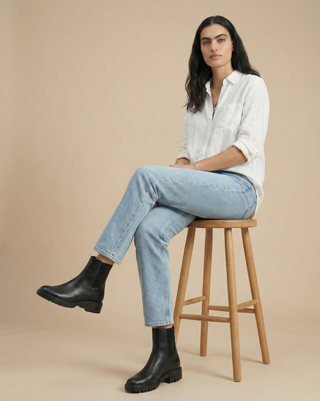 Woman in white shirt, light blue jeans, black Chelsea boots sitting on wooden stool