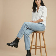Woman in white shirt, light blue jeans, black Chelsea boots sitting on wooden stool