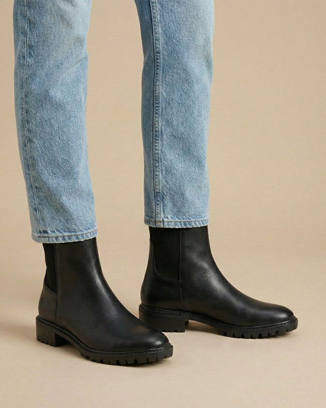 Black leather Chelsea boots with chunky soles worn with light blue jeans on beige background