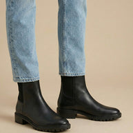 Black leather Chelsea boots with chunky soles worn with light blue jeans on beige background