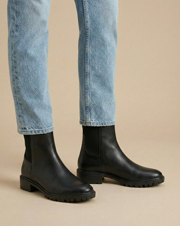 Black leather Chelsea boots with chunky soles worn with light blue jeans on beige background