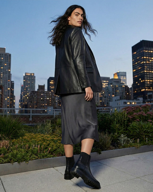 Woman in black leather jacket, satin midi skirt, and boots standing on rooftop at dusk