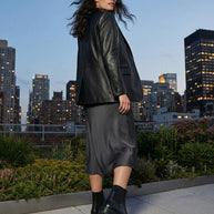 Woman in black leather jacket, satin midi skirt, and boots standing on rooftop at dusk