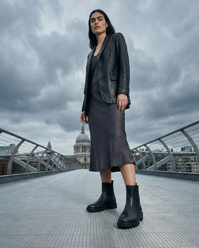 Woman in black leather jacket, dress, and chunky black boots on city bridge with cloudy sky