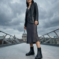 Woman in black leather jacket, dress, and chunky black boots on city bridge with cloudy sky