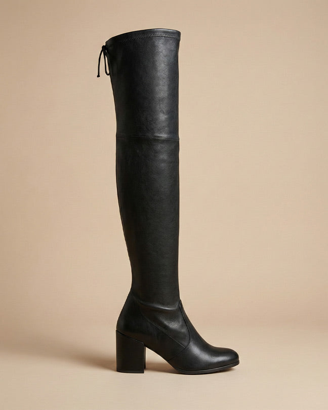 Black over-the-knee high heel boot with block heel and tie detail on beige background