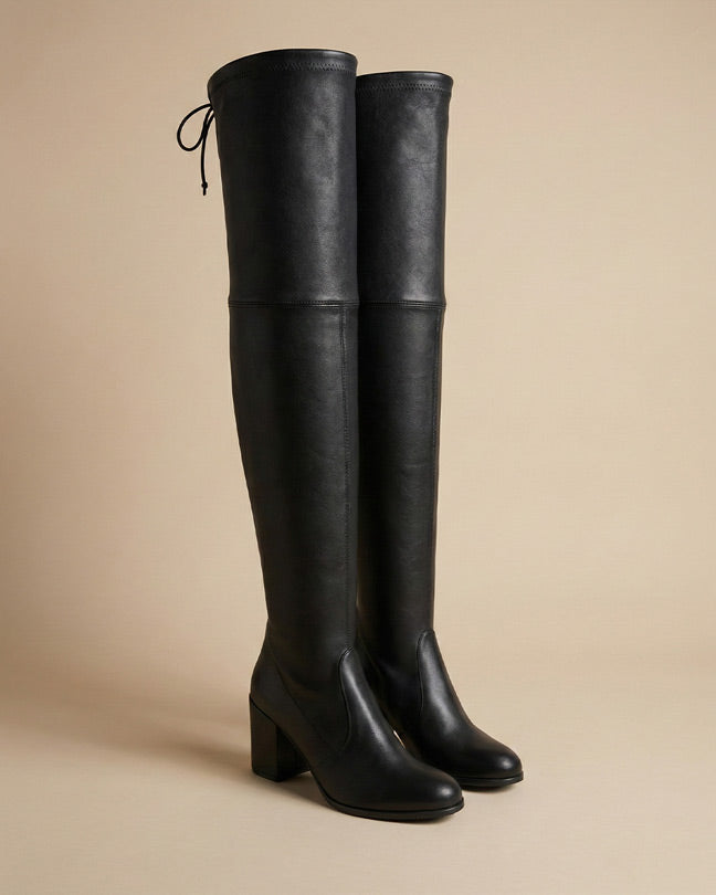 Black over-the-knee block heel boots with tie detail on neutral background