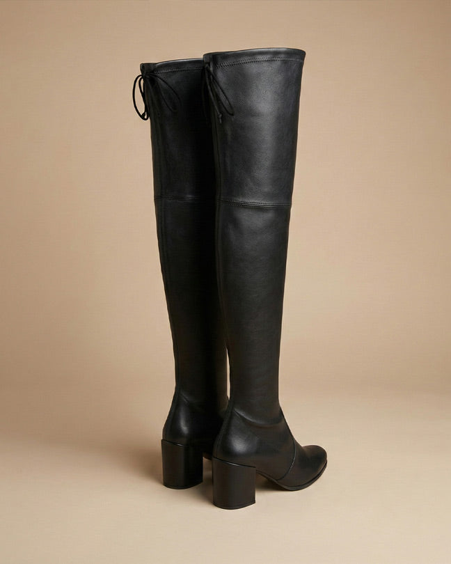 Black over-the-knee heeled leather boots with block heel and tie detail, beige background