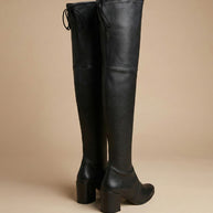 Black over-the-knee heeled leather boots with block heel and tie detail, beige background