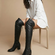 Woman in white shirt dress wearing black over-the-knee heeled leather boots, beige background
