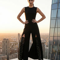 Woman in black sleeveless jumpsuit with thigh-high slits and boots, standing on rooftop at sunset