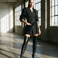 Woman in a black belted blazer dress and over-the-knee black boots in a modern loft