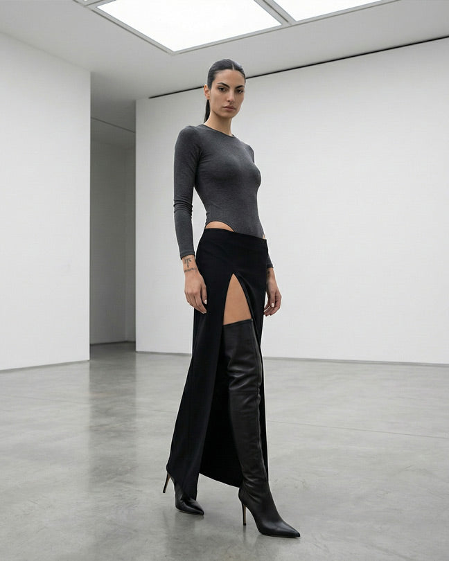 Woman in modern minimalist room wearing black high-slit skirt, grey top, black boots