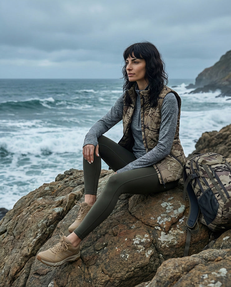 Woman in camouflage vest and hiking gear sitting on rocky coast with backpack by the ocean