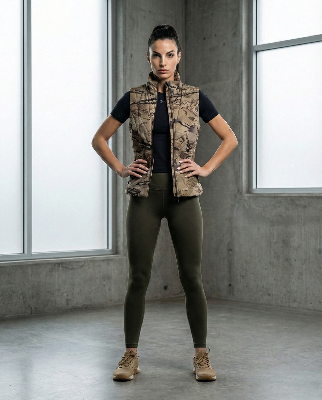 Woman in camouflage vest, black shirt, olive leggings, and tan shoes standing indoors