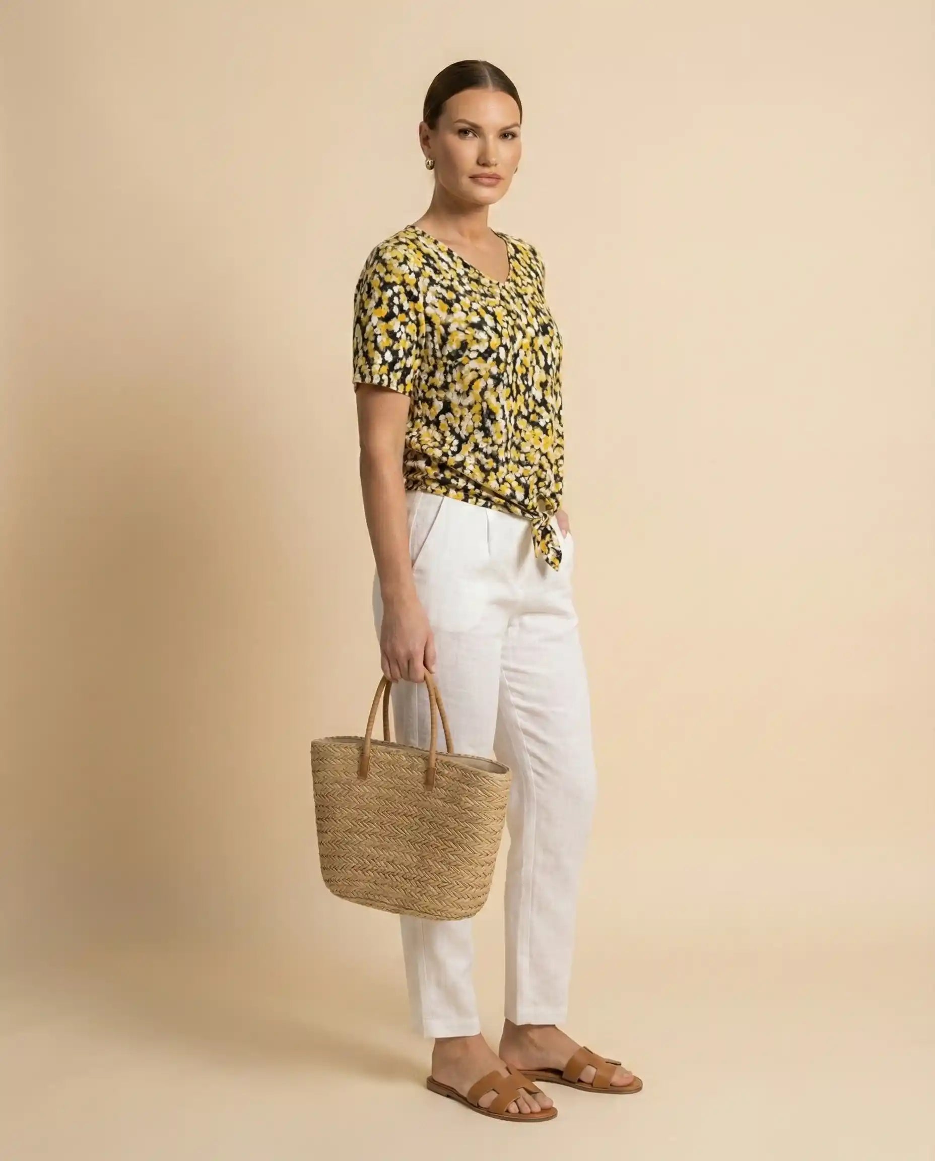 Woman wearing yellow patterned blouse, white pants, carrying straw tote bag and sandals