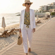 Woman in white linen suit and hat, wearing a yellow spotted top, walking on a boardwalk by the beach