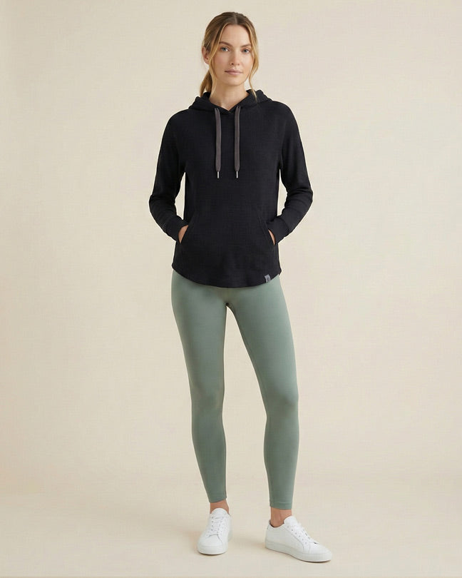Woman in black hoodie, sage green leggings, and white sneakers on neutral background