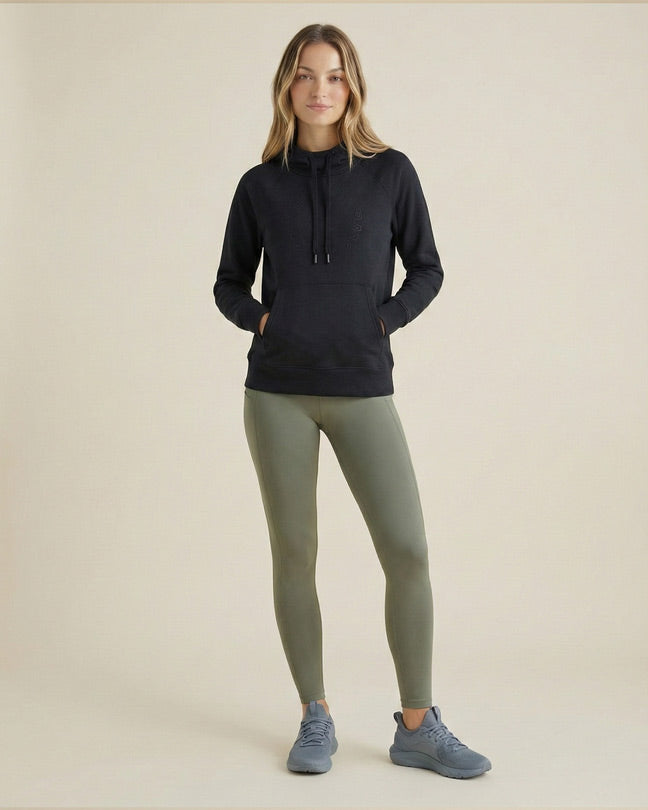 Woman in black hoodie, olive green leggings, and gray sneakers standing in neutral studio