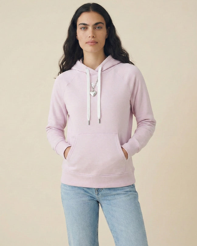 Woman wearing light pink hoodie and blue jeans, hands in pockets, neutral background
