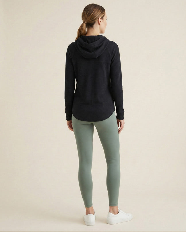 Woman in a black hooded sweatshirt and sage green leggings, standing on a neutral background