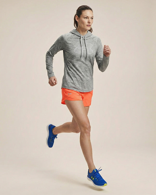 Woman running in gray hoodie, neon orange shorts, and blue athletic shoes against plain background