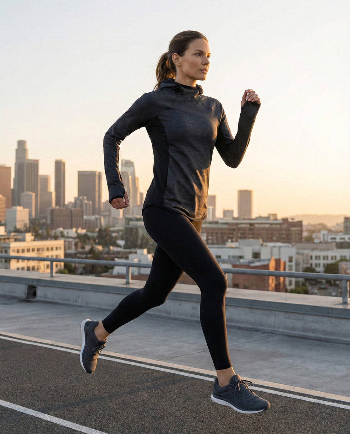 Woman jogging outdoors in grey hoodie and black leggings with city skyline at sunset