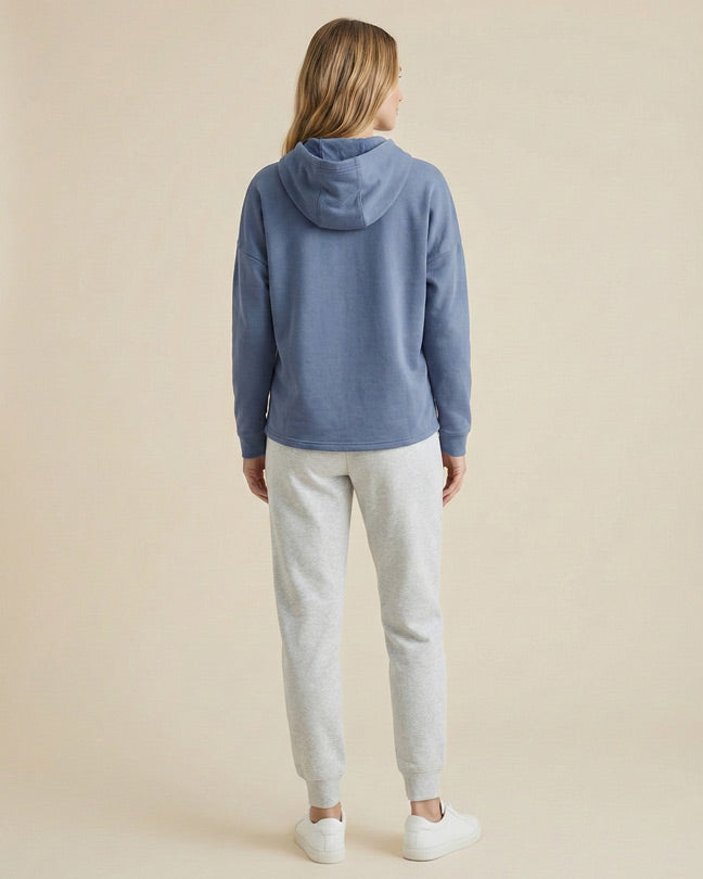 Woman wearing blue hoodie, light gray sweatpants, and white sneakers, standing indoors