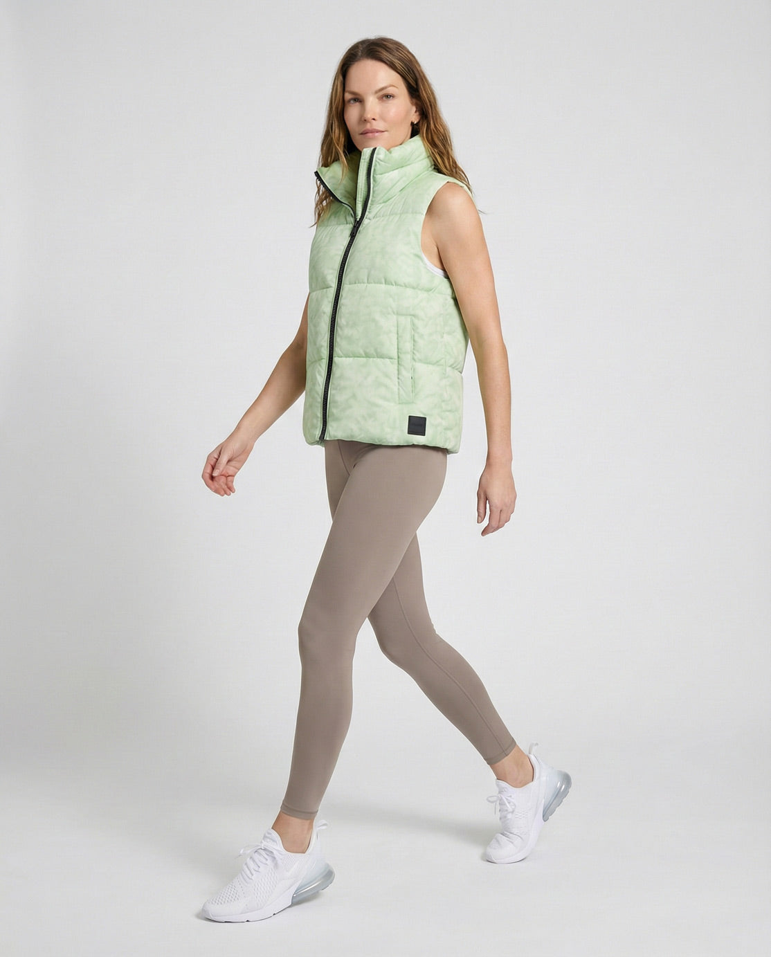 Woman wearing a light green puffer vest, taupe leggings, and white sneakers on a plain background